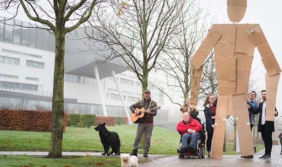 Students outside ҹɫֱ holding a giant card board person up whilst a man with a dog plays guitar