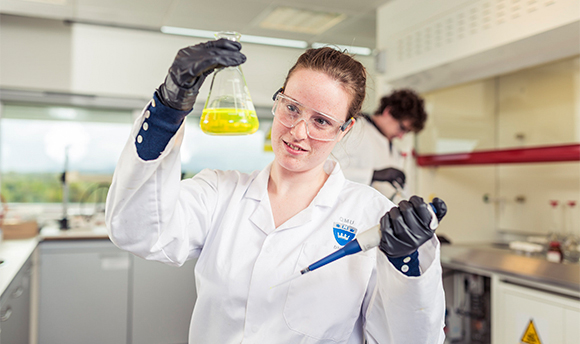 A ҹɫֱ student wearing PPE and holding up a beaker of yellow liquid
